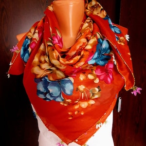 May include: A red floral patterned scarf with a white lace trim and colorful beads. The scarf is draped over a mannequin's neck.