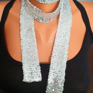 May include: A silver sequin scarf displayed on a mannequin. The scarf is long and narrow, with a shimmering, reflective surface. The sequins are closely spaced, creating a continuous, sparkling effect. The scarf is displayed against a black top.