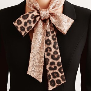May include: A neck scarf featuring a rose gold sequin finish with a black leopard print pattern. The scarf is tied in a bow around the neck and is worn over a solid black blazer with a notched lapel and a deep V-neckline structure.