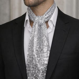May include: A silver sequin ascot is worn over a white collared shirt and under a black suit jacket. The ascot is made of small, reflective sequins. The suit jacket is a classic, tailored style, suitable for formal occasions.