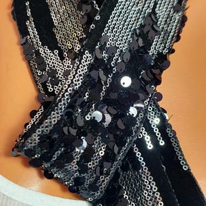 Peut inclure: Une écharpe noire ornée de rangées de paillettes noires brillantes disposées en motif linéaire. L'écharpe est fabriquée à partir d'une base en tissu sombre et est nouée, mettant en valeur la surface réfléchissante des paillettes sur le tissu mat.