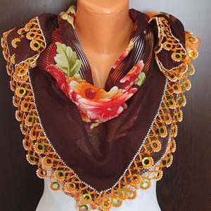 May include: A brown scarf with a floral pattern and a decorative orange and white lace trim. The scarf is draped over a mannequin, showcasing its design and texture. The scarf's floral pattern includes red, orange, and green flowers.