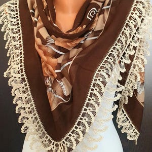 May include: A brown scarf with a floral pattern and a cream-colored lace trim. The scarf is draped over a mannequin, showcasing its design and texture. The lace trim adds a delicate touch to the scarf.