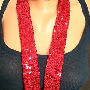 May include: A long, narrow scarf completely covered in small, reflective red sequins. The scarf is wrapped around the neck with two long, flat ends hanging down the front, worn over a plain, dark-colored sleeveless tank top garment.