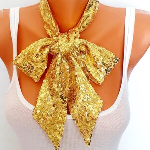 May include: A gold sequin bow tie necklace. The bow tie is made of small, shiny, gold sequins and is tied in a large bow. The necklace is worn around the neck and is a statement piece.