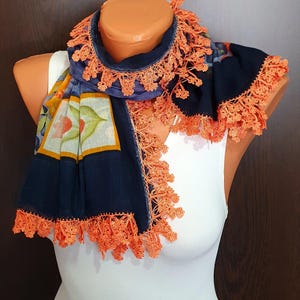 May include: A navy blue scarf with orange lace trim. The scarf features a floral pattern with orange, green, and yellow accents. The scarf is draped over a mannequin.