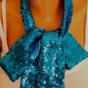May include: A teal sequin neck scarf tied in a bow at the front. The scarf is covered in small, reflective teal sequins that create a shimmering texture. It features a long, narrow band that wraps around the neck and hangs down in two ends.