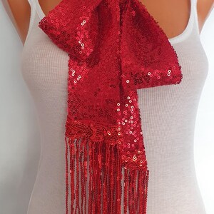 May include: A bright red scarf covered in small, reflective sequins. The scarf features a long, rectangular body that is tied in a knot at the neck, with a decorative braided section at the bottom edge that transitions into long, dangling fringe.