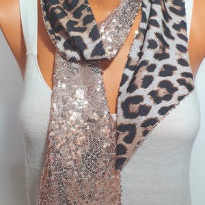 May include: A long scarf featuring a dual-sided design with one side in a brown and black leopard print pattern and the other side covered in shimmering rose gold sequins. The scarf is draped around the neck, showcasing both textures.