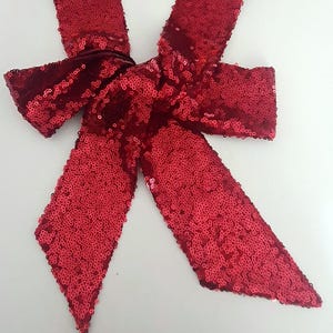 May include: A bright red sequined bow, likely for decoration or embellishment. The bow is tied in the center and has two long, pointed tails. The sequins catch the light, creating a shimmering effect. The bow is on a white background.