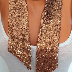 May include: A gold sequin scarf with a V-neck design. The scarf is made of small, shiny sequins and is draped around the neck. The scarf is worn over a white tank top.