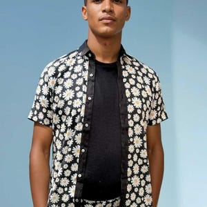 Puede incluir: Conjunto de camisa y pantalón negros, adornados con un estampado de margaritas blancas. La camisa de manga corta está abierta, revelando una camiseta negra debajo. El conjunto tiene cuello y ribete negros.