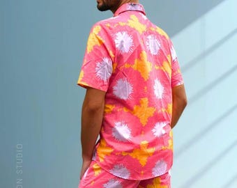 Könnte beinhalten: Ein Set aus einem kurzärmeligen Hemd mit Knopfleiste und passender Shorts aus leuchtend rosa Stoff mit einem sich wiederholenden Muster aus gelben und hellvioletten Sternen. Das Kleidungsstück hat einen Kragen und den Text SEA DRAGON STUDIO.