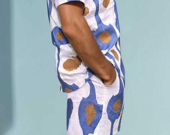 Könnte beinhalten: Ein zweiteiliges Set, bestehend aus einem kurzärmeligen Hemd und Shorts. Das Set ist weiß mit einem sich wiederholenden Muster aus blauen und braunen abstrakten Formen. Die Person trägt das Set mit der Hand in der Tasche.