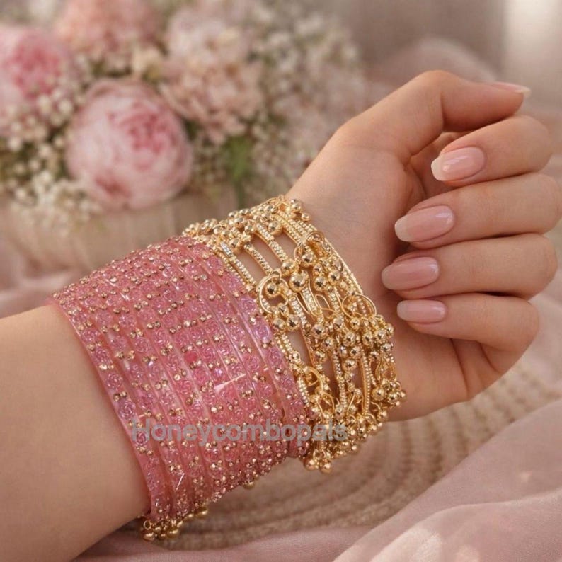 Op de afbeelding: Een pols is versierd met een reeks roze armbanden en een gouden armband. De roze armbanden zijn transparant met gouden details. De gouden armband heeft een sierlijk ontwerp. Roze bloemen zijn zichtbaar op de achtergrond.