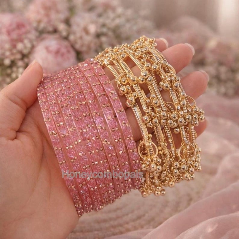 Op de afbeelding: Een hand toont twee sets armbanden. E&eacute;n set is lichtroze met kleine, sprankelende versieringen. De andere set is goudkleurig, met bungelende balbedels. De achtergrond is zachtroze, met een vage bloemstuk.