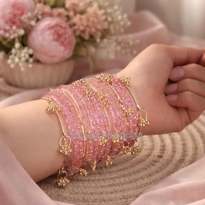 Op de afbeelding: Een verzameling roze armbanden met goudkleurige accenten en bungelende bedels. De armbanden zijn op een pols gestapeld, tegen een zachtroze achtergrond met een boeket roze bloemen op de achtergrond.