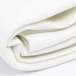 May include: Close-up of a folded stack of white fabric. The fabric appears to be a soft, textured material, possibly cotton or linen. The folds create a layered effect, with the edges of the fabric curving gently. The overall impression is one of softness and purity.
