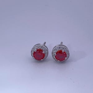 May include: A pair of silver stud earrings with a red gemstone surrounded by a halo of smaller white gemstones.