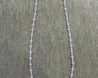 May include: A silver-toned necklace with a repeating starburst design. The necklace is composed of small, sparkling stones set in a linear pattern. The chain is displayed against a neutral, textured background.