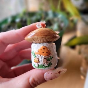 May include: A small ceramic mushroom-shaped trinket box with a lid. The box is painted white with brown and orange details. The lid features a brown mushroom cap with white spots. The box is decorated with green leaves and small mushrooms.