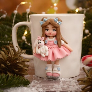 May include: A white ceramic mug with a handmade polymer clay doll attached. The doll is wearing a pink dress and holding a white bunny. The doll has brown hair with blue bows. The background includes pine needles and fairy lights.