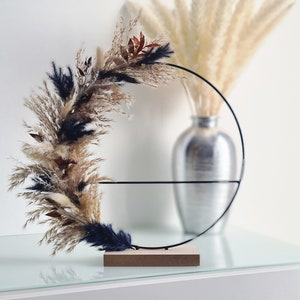 Peut inclure: Un cercle en métal noir avec une base en bois, décoré d'herbe de pampa séchée dans des tons beige, brun et bleu. Le cercle est posé sur une surface en verre.
