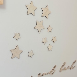 Può includere: Decorazioni da parete a forma di stelle in legno di varie dimensioni. La frase "moon and back" è scritta in corsivo sotto le stelle. Le stelle e il testo sono in legno chiaro, creando un'atmosfera minimalista.