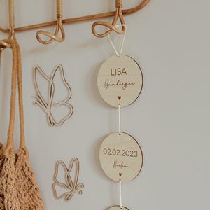 May include: Wooden baby mobile with three round wooden tags. The top tag says "LISA Gumburger". The middle tag says "02.02.2023 Birth". The bottom tag says "14:35 3135 G 55 cm".