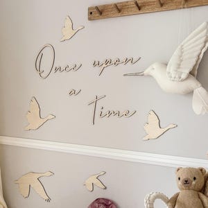 Puede incluir: Decoración mural de guardería con recortes de pájaros de madera y la frase "Once upon a Time" en cursiva. Un colibrí de tela blanca cuelga del techo. Una percha de madera está montada encima del texto. Una cuna y una cesta con un osito de peluche están en primer plano.