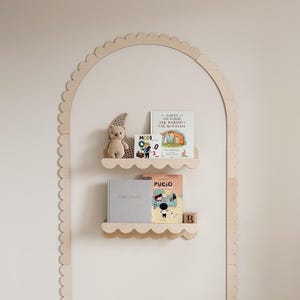 Könnte beinhalten: Ein hölzerner, wellenförmiger Bogen umrahmt zwei Regale mit Kinderbüchern und einem Stofftier. Die Regale und der Bogen sind aus hellem Holz gefertigt. Die Bücher haben farbenfrohe Einbände und Text. Die Wand ist in einer neutralen Farbe gestrichen.