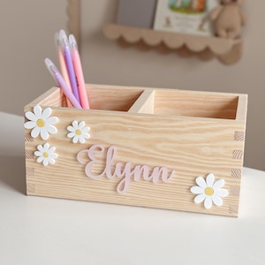 Puede incluir: Un organizador de escritorio de madera clara con dos compartimentos, decorado con acentos de margaritas blancas y el nombre "Elynn" en escritura rosa. El organizador contiene varios bolígrafos y lápices. Una estantería a juego es visible.