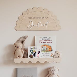 Puede incluir: Estanterías de madera para guardería con bordes festoneados, que sostienen libros, un oso de peluche y objetos decorativos. Un letrero de madera en la parte superior dice "KLEINE BIBLIOTHEK VON Juliet". Las estanterías están montadas en una pared clara.