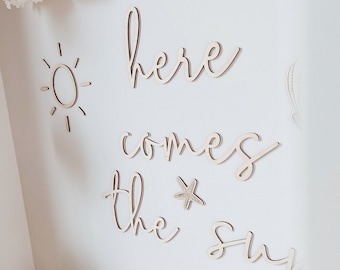 Può includere: Decorazione da parete in legno con le parole "here comes the sun" in un font corsivo. La decorazione include anche un sole, una stella marina, una conchiglia e una tartaruga.