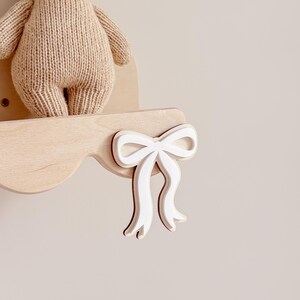 Può includere: Un peluche lavorato a maglia color beige chiaro a forma di coniglio è appoggiato su una mensola da parete in legno chiaro. La mensola presenta un ritaglio decorativo a forma di fiocco bianco fissato sul bordo anteriore del legno.