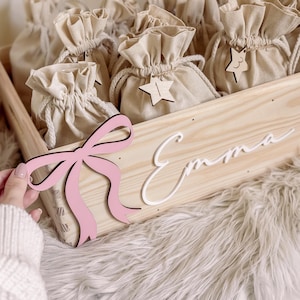 Può includere: Una cassetta in legno chiaro con un fiocco in legno rosa e il nome Emma in corsivo bianco. All'interno della cassetta ci sono diversi sacchetti in tessuto beige con coulisse, ognuno decorato con una piccola stella in legno numerata.