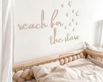 May include: A wooden crib with a white and beige bedding set. The crib has a braided rope bumper and a stuffed animal toy. The wall behind the crib has a wooden sign that reads "reach for the stars" with wooden stars surrounding it.