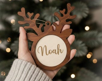 May include: A wooden Christmas ornament in the shape of a reindeer head. The ornament has a circular center with the name "Noah" engraved. The antlers and outer frame are a dark brown, while the center is a lighter wood color. A gold string is attached for hanging.