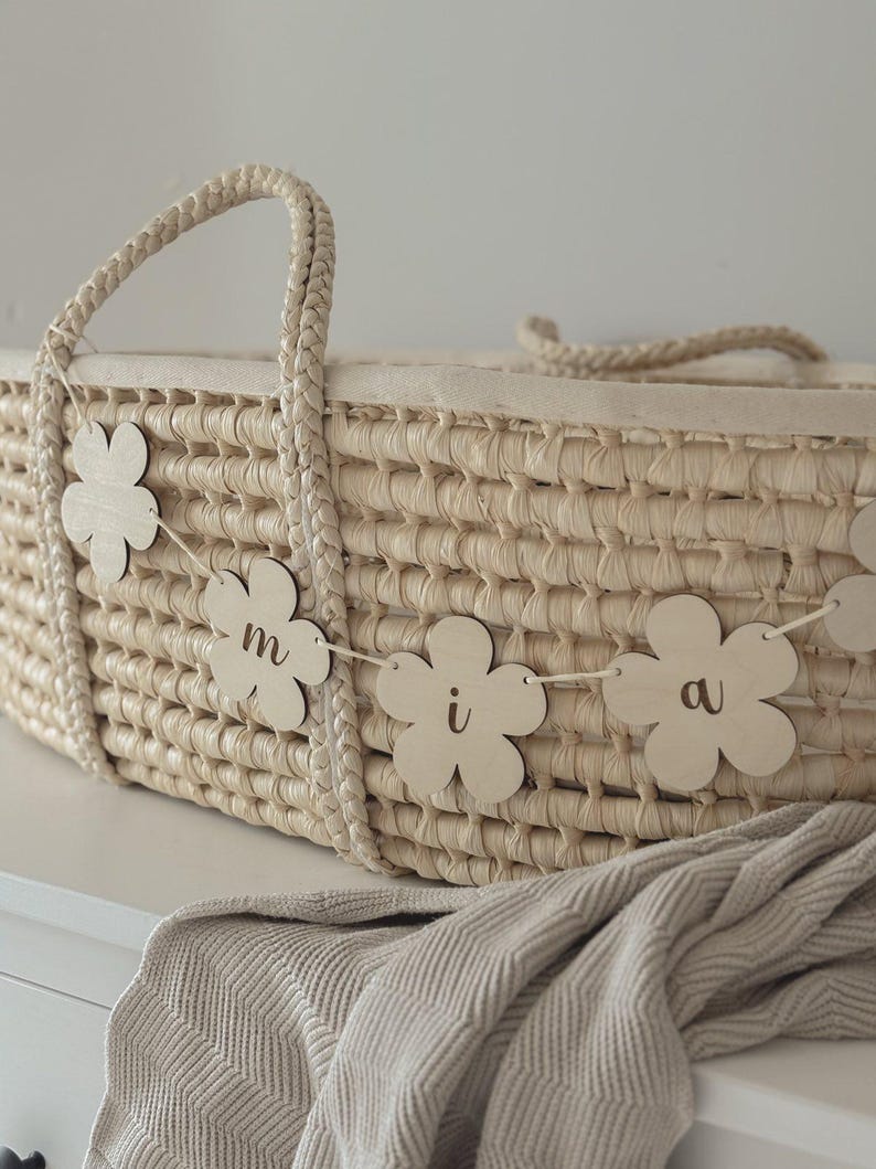 May include: A woven wicker bassinet with a braided handle and a soft, light-colored interior. A garland of wooden flower cutouts with the letters "m i a" hangs across the front. A beige blanket is draped over the edge.