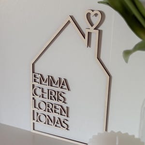 May include: Wooden house-shaped wall decor with a heart on top and the names Emma, Chris, Loren, and Jonas written in a modern font.