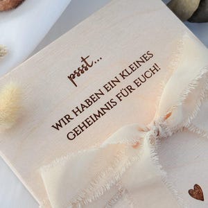 May include: A light-colored wooden box with the text "pssst... WIR HABEN EIN KLEINES GEHEIMNIS F&Uuml;R EUCH!" in brown lettering. The box is tied with a cream-colored ribbon and has a small heart etched on the side.