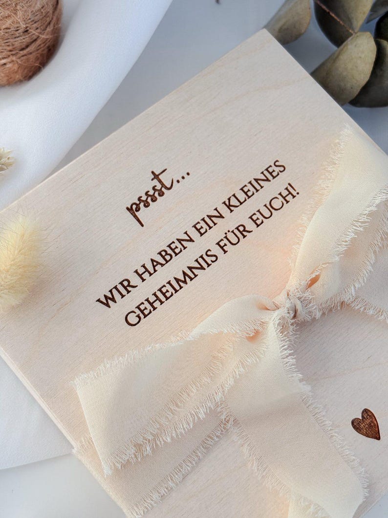 May include: A light-colored wooden box with the text "pssst... WIR HABEN EIN KLEINES GEHEIMNIS F&Uuml;R EUCH!" in brown lettering. The box is tied with a cream-colored ribbon and has a small heart etched on the side.