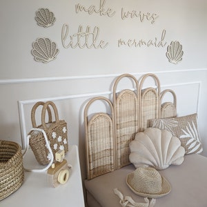 May include: A coastal-themed bedroom scene with a light beige color palette. The wall features wooden seashell decorations and the phrase "make waves little mermaid." A woven headboard, shell-shaped pillow, and straw accessories complete the decor.