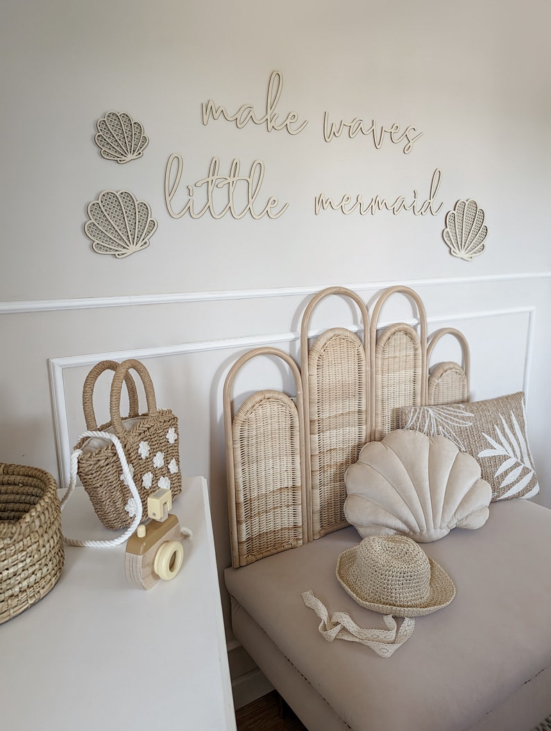 May include: A coastal-themed bedroom scene with a light beige color palette. The wall features wooden seashell decorations and the phrase "make waves little mermaid." A woven headboard, shell-shaped pillow, and straw accessories complete the decor.
