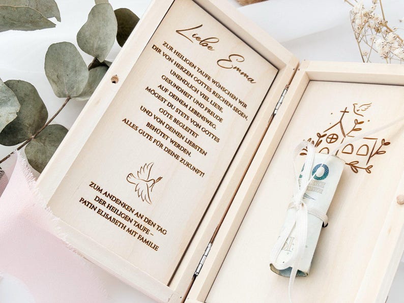 May include: A wooden box with a dove and church design engraved on the lid. The box is open and contains a white envelope with a rolled up bill of money inside. The box also has a message inside that reads "Liebe Emma Zur heiligen Taufe w&uuml;nschen wir dir von Herzen Gottes reichen Segen, unendlich viel Liebe. Gesundheit und Freude auf deinem Lebensweg. M&ouml;gest du stets von Gottes gute begleitet und von deinen Liebsten beh&uuml;tet werden. Alles Gute f&uuml;r deine Zukunft! Zum Andenken an den Tag der heiligen Taufe - Patin Elisabeth mit Familie"