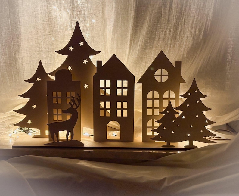 Puede incluir: Escena de pueblo navide&ntilde;o de madera con casas, &aacute;rboles y una silueta de reno recortados. Los &aacute;rboles tienen recortes de estrellas y las casas tienen recortes de ventanas. La escena est&aacute; iluminada desde atr&aacute;s, creando un brillo c&aacute;lido. Una decoraci&oacute;n festiva.