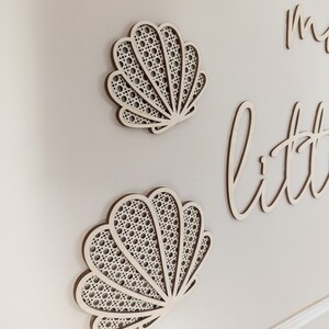 May include: Two wooden seashell wall decorations with a woven pattern, mounted on a light-colored wall. The shells are light brown with a woven, off-white interior. The word "little" is visible in cursive script.