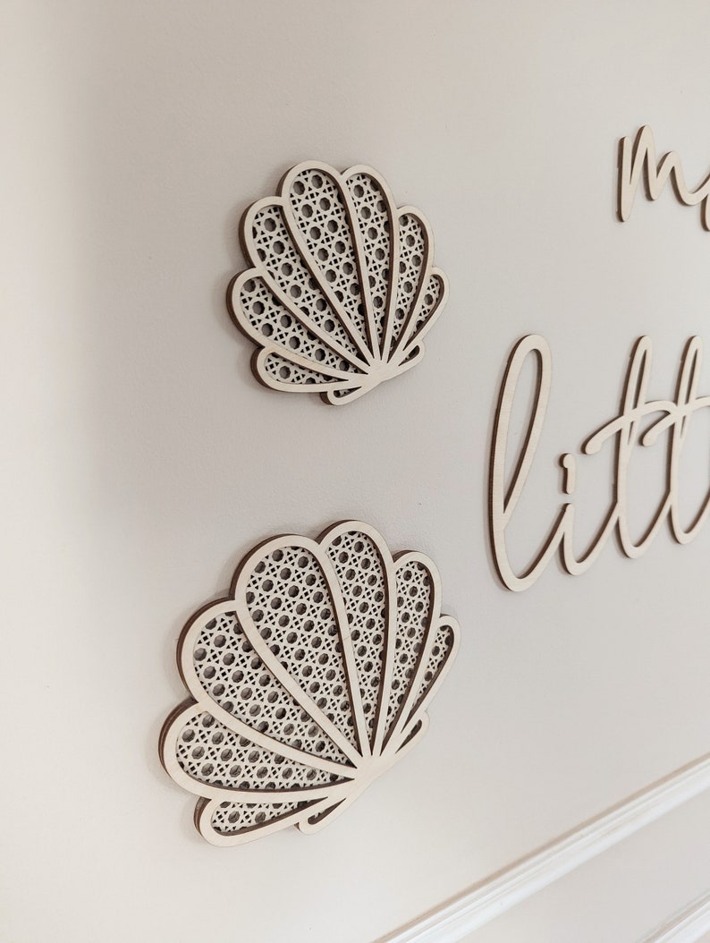 May include: Two wooden seashell wall decorations with a woven pattern, mounted on a light-colored wall. The shells are light brown with a woven, off-white interior. The word "little" is visible in cursive script.