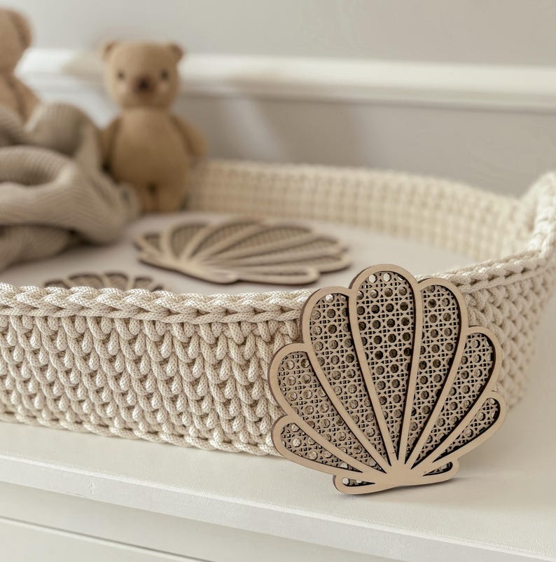 May include: A cream-colored, hand-crocheted basket with a seashell-shaped wooden accent. The basket has a sturdy, woven texture and is placed on a white surface. Two small teddy bears and a blanket are visible in the background.
