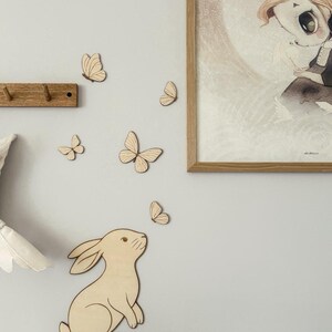 May include: Wooden wall decor featuring a light brown rabbit looking up at a group of wooden butterflies. A framed illustration with a child-like figure is on the wall. A wooden coat rack is also visible.